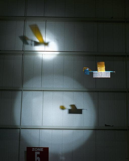 NASA image: An experimental radio-controlled model aircraft casts two unique shadows as it flies inside a Dryden hangar using two spotlights as energy sources.