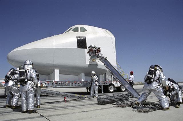 NASA image: While rescue personnel prepare evacuation litters, two stand-in "astronauts" prepare to use an exit slide from a Shuttle mockup during a training exercise