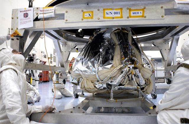 NASA image: KENNEDY SPACE CENTER, FLA. - Workers in the Payload Hazardous Servicing Facility lower the backshell over the Mars Exploration Rover 1 (MER-1).  The backshell is a protective cover for the rover.  NASA's twin Mars Exploration Rovers are designed to study the history of water on Mars. These robotic geologists are equipped with a robotic arm, a drilling tool, three spectrometers, and four pairs of cameras that allow them to have a human-like, 3D view of the terrain. Each rover could travel as far as 100 meters in one day to act as Mars scientists' eyes and hands, exploring an environment where humans can't yet go.  MER-1 is scheduled to launch June 25 as MER-B aboard a Delta II rocket from Cape Canaveral Air Force Station.