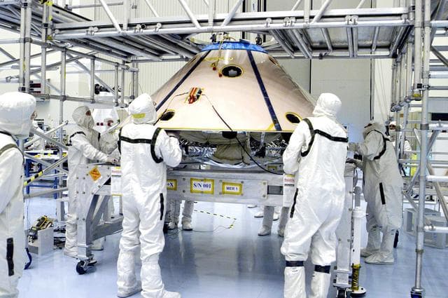 NASA image: KENNEDY SPACE CENTER, FLA. -  Workers watch as an overhead crane begins to lift the backshell with the Mars Exploration Rover 1 (MER-1) inside.  The backshell will be moved and attached to the lower heat shield.  NASA's twin Mars Exploration Rovers are designed to study the history of water on Mars. These robotic geologists are equipped with a robotic arm, a drilling tool, three spectrometers, and four pairs of cameras that allow them to have a human-like, 3D view of the terrain. Each rover could travel as far as 100 meters in one day to act as Mars scientists' eyes and hands, exploring an environment where humans can't yet go.  MER-1 is scheduled to launch June 25 as MER-B aboard a Delta II rocket from Cape Canaveral Air Force Station.