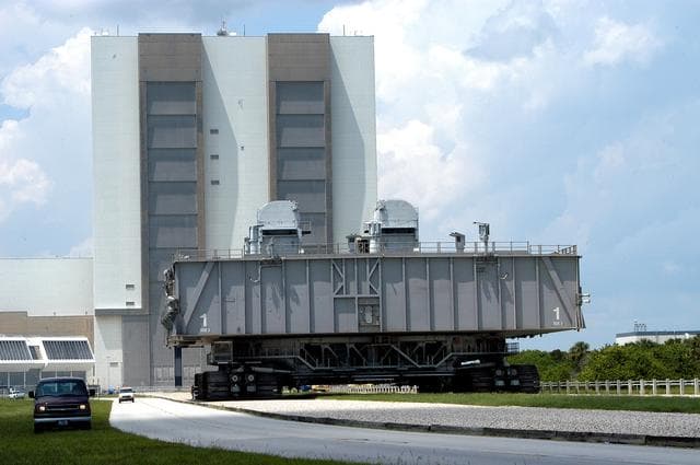 NASA image: KENNEDY SPACE CENTER, FLA.  -  Crawler-transporter (CT) number 2, moves away from the Vehicle Assembly Building with a Mobile Launcher Platform (MLP) on top on a test run to the launch pad.  The CT recently underwent modifications to the cab.  The CT moves Space Shuttle vehicles, situated on the MLP, between the VAB and launch pad.  Moving on four double-tracked crawlers, the CT uses a laser guidance system and a leveling system for the journey that keeps the top of a Space Shuttle vertical within plus- or minus-10 minutes of arc.  The system enables the CT-MLP-Shuttle to negotiate the ramp leading to the launch pads and keep the load level.  Unloaded, the CT weighs 6 million pounds.  Seen on top of the MLP are two tail service masts that support the fluid, gas and electrical requirements of the orbiter’s liquid oxygen and liquid hydrogen aft umbilicals.