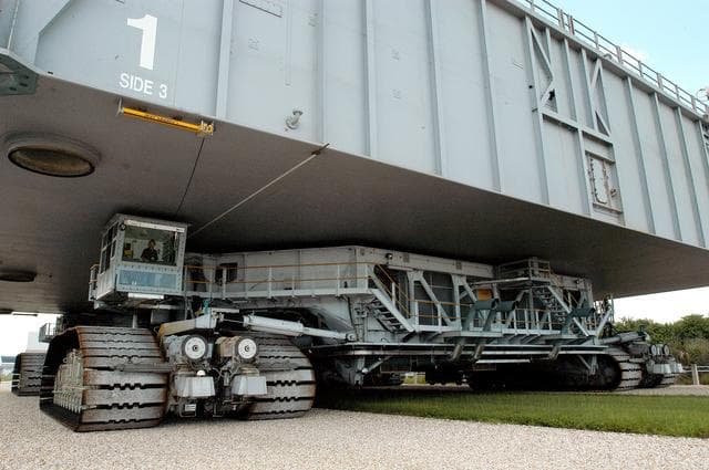 NASA image: KENNEDY SPACE CENTER, FLA.  -  A closeup of crawler-transporter (CT) number 2 shows the cab (left, above the tracks) that recently underwent modifications.  The CT is transporting a Mobile Launch Platform (MLP) on a test run to the pad.  The CT moves Space Shuttle vehicles, situated on the MLP, between the VAB and launch pad.  Moving on four double-tracked crawlers, the CT uses a laser guidance system and a leveling system for the journey that keeps the top of a Space Shuttle vertical within plus- or minus-10 minutes of arc.  The system enables the CT-MLP-Shuttle to negotiate the ramp leading to the launch pads and keep the load level.  Unloaded, the CT weighs 6 million pounds.  Seen on top of the MLP are two tail service masts that support the fluid, gas and electrical requirements of the orbiter’s liquid oxygen and liquid hydrogen aft umbilicals.