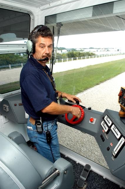 NASA image: KENNEDY SPACE CENTER, FLA.  -  Inside the cab of crawler-transporter (CT) number 2, driver Sam Dove, with United Space Alliance, operates the vehicle on a test run to the launch pad.  The CT recently underwent modifications to the cab.   The CT is transporting a Mobile Launch Platform (MLP).  The CT moves Space Shuttle vehicles, situated on the MLP, between the VAB and launch pad.  Moving on four double-tracked crawlers, the CT uses a laser guidance system and a leveling system for the journey that keeps the top of a Space Shuttle vertical within plus- or minus-10 minutes of arc.  The system enables the CT-MLP-Shuttle to negotiate the ramp leading to the launch pads and keep the load level.  Unloaded, the CT weighs 6 million pounds.  Seen on top of the MLP are two tail service masts that support the fluid, gas and electrical requirements of the orbiter’s liquid oxygen and liquid hydrogen aft umbilicals.