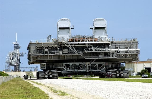 NASA image: KENNEDY SPACE CENTER, FLA.  -   Crawler-transporter (CT) number 2 nears the launch pad with a Mobile Launcher Platform (MLP) on top.  After recent modifications to the cab and muffler system, the CT was taken on a test run.   The CT moves Space Shuttle vehicles, situated on the MLP, between the VAB and launch pad.  Moving on four double-tracked crawlers, the CT uses a laser guidance system and a leveling system for the journey that keeps the top of a Space Shuttle vertical within plus- or minus-10 minutes of arc.  The system enables the CT-MLP-Shuttle to negotiate the ramp leading to the launch pads and keep the load level.  Unloaded, the CT weighs 6 million pounds.  Seen on top of the MLP are two tail service masts that support the fluid, gas and electrical requirements of the orbiter’s liquid oxygen and liquid hydrogen aft umbilicals.