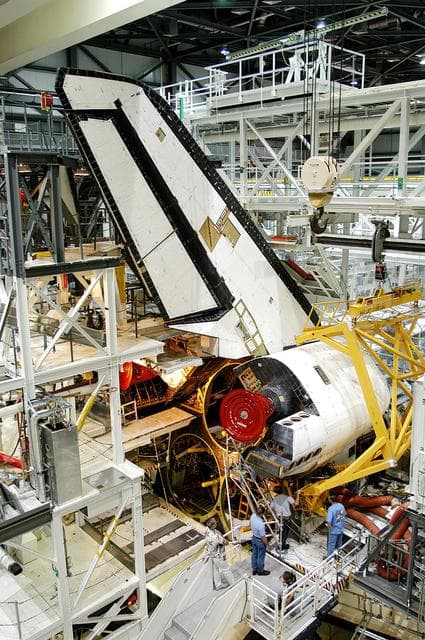 NASA image: KENNEDY SPACE CENTER, FLA. -  Workers in the Orbiter Processing Facility prepare to remove one of two orbital maneuvering system (OMS) pods from Endeavour.  The OMS pods are attached to the upper aft fuselage left and right sides.  Fabricated primarily of graphite epoxy composite and aluminum, each pod is 21.8 feet long and 11.37 feet wide at its aft end and 8.41 feet wide at its forward end, with a surface area of approximately 435 square feet. Each pod houses the Reaction Control System propulsion components used for inflight maneuvering and is attached to the aft fuselage with 11 bolts.  OMS pods are removed during Orbiter Major Modifications. Once removed, the OMS pods undergo in-depth structural inspections, system checks and the thrusters are changed out.