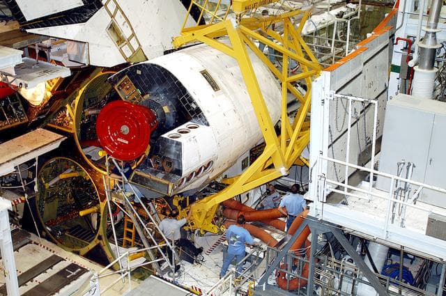 NASA image: KENNEDY SPACE CENTER, FLA. -  Workers in the Orbiter Processing Facility prepare to remove one of two orbital maneuvering system (OMS) pods from Endeavour.  The OMS pods are attached to the upper aft fuselage left and right sides.  Fabricated primarily of graphite epoxy composite and aluminum, each pod is 21.8 feet long and 11.37 feet wide at its aft end and 8.41 feet wide at its forward end, with a surface area of approximately 435 square feet. Each pod houses the Reaction Control System propulsion components used for inflight maneuvering and is attached to the aft fuselage with 11 bolts.   OMS pods are removed during Orbiter Major Modifications. Once removed, the OMS pods undergo in-depth structural inspections, system checks and the thrusters are changed out.