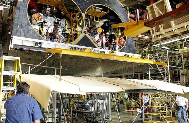 NASA image: KENNEDY SPACE CENTER, FLA. - In the Orbiter Processing Facility, workers look down from spaces allotted for the main engines as the rear body flap is lifted for installation on the orbiter Discovery.  The body flap, which is temporarily under protective covering, attaches below the main engines.