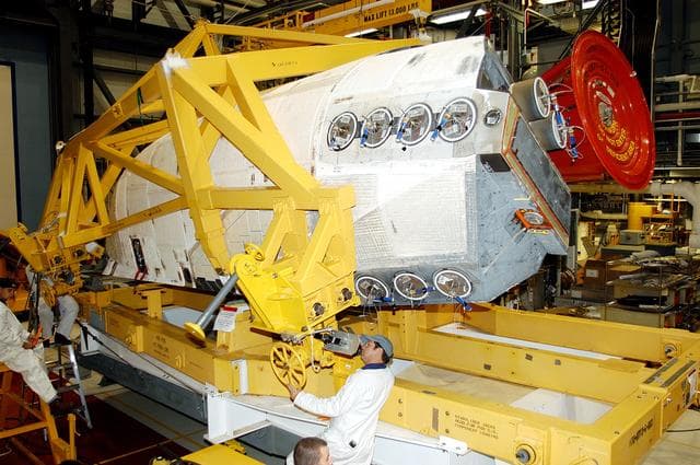 NASA image: KENNEDY SPACE CENTER, FLA. - In the Orbiter Processing Facility, one of two orbital maneuvering system (OMS) pods is being moved for installation on Atlantis.  The OMS pods are attached to the upper aft fuselage left and right sides. Fabricated primarily of graphite epoxy composite and aluminum, each pod is 21.8 feet long and 11.37 feet wide at its aft end and 8.41 feet wide at its forward end, with a surface area of approximately 435 square feet. Each pod houses the Reaction Control System propulsion components used for inflight maneuvering and is attached to the aft fuselage with 11 bolts.