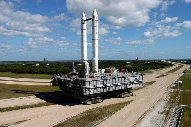 NASA image: KENNEDY SPACE CENTER, FLA. -  Mobile Launcher Platform (MLP) number 3 and a set of twin solid rocket boosters bolted to it, atop the crawler-transporter, crawl to the intersection in the crawlerway in support of the second engineering analysis vibration test on the crawler and MLP.  In the background are Launch Pads 39A (right) and 39B (left).  The crawler is moving at various speeds up to 1 mph in an effort to achieve vibration data gathering goals as it leaves the VAB, travels toward Launch Pad 39A and then returns.  The boosters are braced at the top for stability.  The primary purpose of these rollout tests is to gather data to develop future maintenance requirements on the transport equipment and the flight hardware. Various parts of the MLP and crawler transporter have been instrumented with vibration data collection equipment.