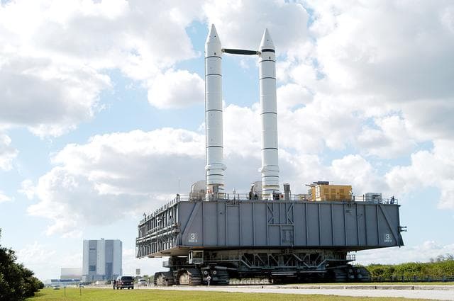 NASA image: KENNEDY SPACE CENTER, FLA. - The crawler-transporter carrying Mobile Launcher Platform (MLP) number 3, with a set of twin solid rocket boosters bolted atop, crawls to the intersection in the crawlerway in support of the second engineering analysis vibration test on the crawler and MLP.  From this perspective, the Launch Control Center (left) and the 525-foot-tall Vehicle Assembly Building (right) in the background appear dwarfed by the 184-foot-tall boosters. The crawler is moving at various speeds up to 1 mph in an effort to achieve vibration data gathering goals as it leaves the VAB, travels toward Launch Pad 39A and then returns. The boosters are braced at the top for stability. The primary purpose of these rollout tests is to gather data to develop future maintenance requirements on the transport equipment and the flight hardware. Various parts of the MLP and crawler transporter have been instrumented with vibration data collection equipment.