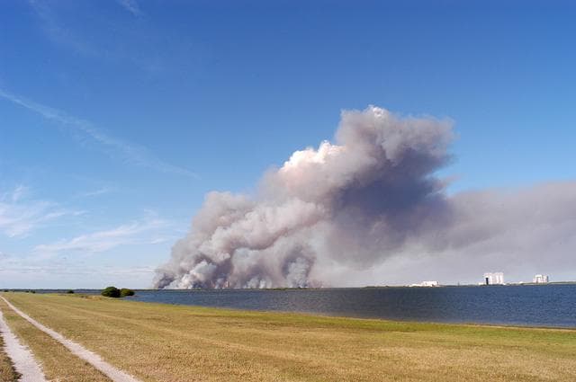 NASA image: KENNEDY SPACE CENTER, FLA. - A successful controlled burn near KSC’s Launch Complex 39 area creates clouds of smoke in a clear blue sky.  The water seen is the Banana River.