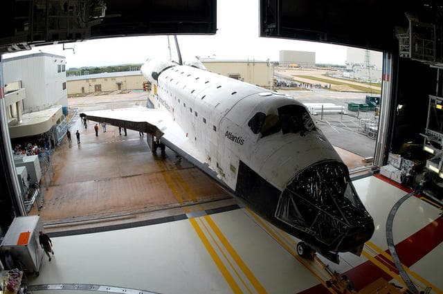 NASA image: KENNEDY SPACE CENTER, FLA. - The Space Shuttle orbiter Atlantis backs out of the Orbiter Processing Facility (OPF) for its move to the Vehicle Assembly Building (VAB). The move will allow work to be performed in the OPF that can only be accomplished while the bay is empty. Work scheduled in the processing facility includes annual validation of the bay's cranes, work platforms, lifting mechanisms, and jack stands. Atlantis will remain in the VAB for about 10 days, then return to the OPF as work resumes to prepare it for launch in September 2004 on the first return-to-flight mission, STS-114.