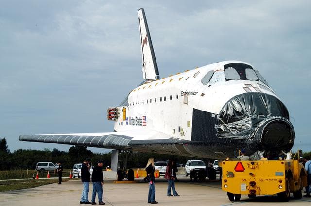 NASA image: KENNEDY SPACE CENTER, FLA. -- After Endeavour’s rollout from inside the Orbiter Processing Facility, the transporter (foreground) prepares to tow it to the Vehicle Assembly Building for temporary transfer.  A protective cover surrounds the nose of Endeavour.  The move to the VAB allows work to be performed in the OPF that can only be accomplished while the bay is empty. Work scheduled in the OPF includes annual validation of the bay’s cranes, work platforms, lifting mechanisms and jack stands. Endeavour will remain in the VAB for approximately 12 days, then return to the OPF.