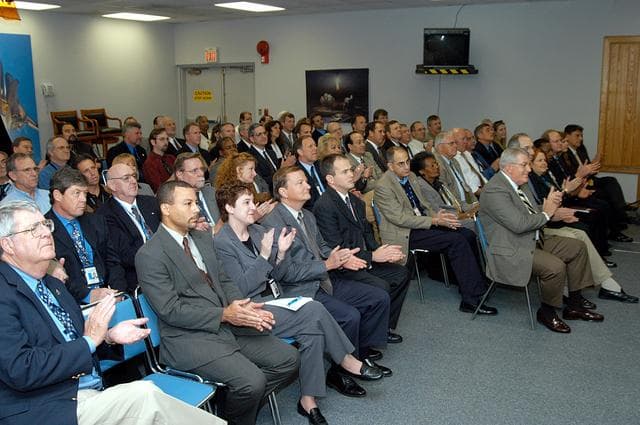 NASA image: KENNEDY SPACE CENTER, FLA. -- From the KSC television studio, KSC management and other employees applaud President George W. Bush, who addressed the public and an assembly of government officials at NASA Headquarters, outlining a new focus and vision for the space agency.  Fourth from left is Mike Leinbach, Shuttle launch director; at right, front row, are Bill Pickavance vice president and associate program manager of Florida Operations, United Space Alliance (USA)  and Howard DeCastro, USA vice president and Space Shuttle program manager.  The President stated his goals for NASA’s new mission: Completing the International Space Station, retiring the Space Shuttle orbiters, developing a new crew exploration vehicle, and returning to the moon and beyond within the next two decades.   Pres. Bush was welcomed by NASA Administrator Sean O’Keefe and Expedition 8 Commander Michael Foale, who greeted him from the International Space Station.  Members of the Washington, D.C., audience included astronauts Eileen Collins, Ed Lu and Michael Lopez-Alegria, and former astronaut Gene Cernan.