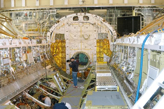 NASA image: KENNEDY SPACE CENTER, FLA. -  In the Orbiter Processing Facility, a cameraman films part of Discovery’s payload bay for a special feature on the KSC Web.  In the background is the open hatch of the airlock, located inside the middeck of the spacecraft’s pressurized crew cabin. The airlock is sized to accommodate two fully suited flight crew members simultaneously. Support functions include airlock depressurization and repressurization, extravehicular activity equipment recharge, liquid-cooled garment water cooling, EVA equipment checkout, donning and communications. The outer hatch isolates the airlock from the unpressurized payload bay when closed and permits the EVA crew members to exit from the airlock to the payload bay when open.