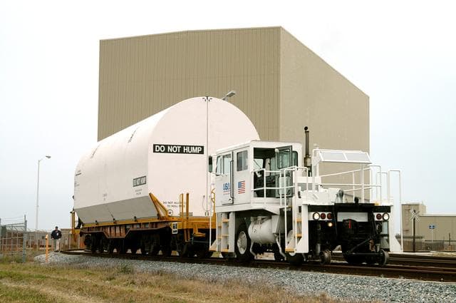 NASA image: KENNEDY SPACE CENTER, FLA. - An engine pulls the container enclosing a segment of a solid rocket booster from the Rotation Processing and Surge Facility.  The container will join others on the main track for a trip to Utah where the segments will undergo firing.  The segments were part of the STS-114 stack.  It is the first time actual flight segments that had been stacked for flight in the VAB are being returned for testing.  They will undergo firing, which will enable inspectors to check the viability of the solid and verify the life expectancy for stacked segments.
