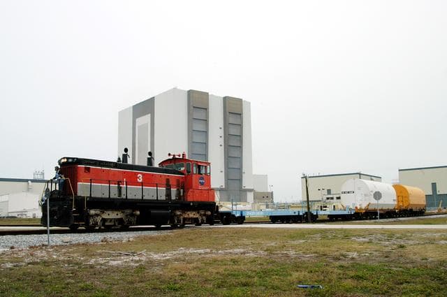 NASA image: KENNEDY SPACE CENTER, FLA. - The red NASA engine moves forward past the Vehicle Assembly Building with its cargo of containers enclosing segments of a solid rocket booster being returned to Utah for testing.  The segments were part of the STS-114 stack.  It is the first time actual flight segments that had been stacked for flight in the VAB are being returned for testing.  They will undergo firing, which will enable inspectors to check the viability of the solid and verify the life expectancy for stacked segments.