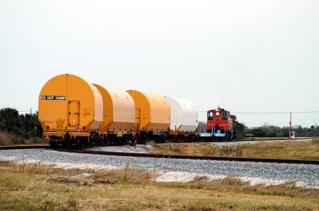 NASA image: KENNEDY SPACE CENTER, FLA. - The red NASA engine hauls its cargo toward Titusville, Fla. The containers enclose segments of a solid rocket booster being returned to Utah for testing.  The segments were part of the STS-114 stack.  It is the first time actual flight segments that had been stacked for flight in the VAB are being returned for testing.  They will undergo firing, which will enable inspectors to check the viability of the solid and verify the life expectancy for stacked segments.