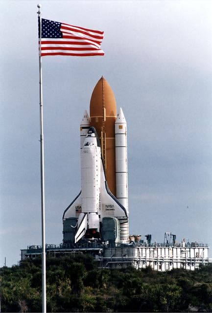 NASA image: KENNEDY SPACE CENTER, FLA. -  The Space Shuttle Discovery on its Mobile Launcher Platform makes its slow 3.4-mile trek from the Vehicle Assembly Building to Launch Pad 39A in preparation for the STS-82 mission.  In the foreground is the U.S. flag at the Press Site area.  A seven-member crew will perform the second servicing of the orbiting Hubble Space Telescope (HST) during the 10-day STS-82 mission, which is targeted for a Feb. 11 liftoff.