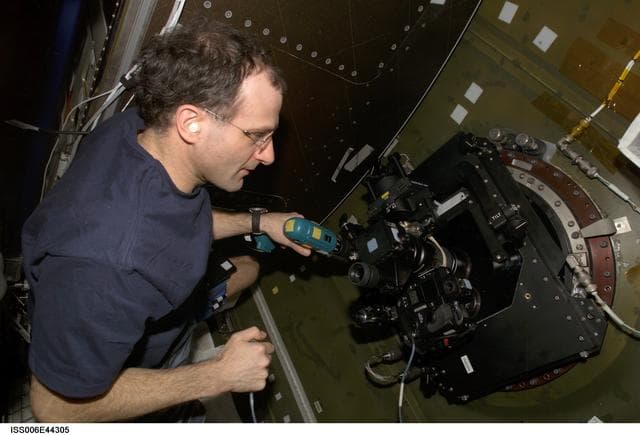 NASA image: Pettit runs a drill while looking through  a camera mounted on the Nadir window in the U.S. Lab