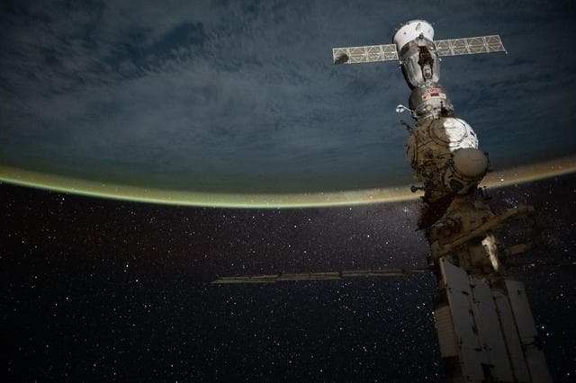NASA image: A yellow-green airglow blankets Earth’s horizon beneath a star-filled sky