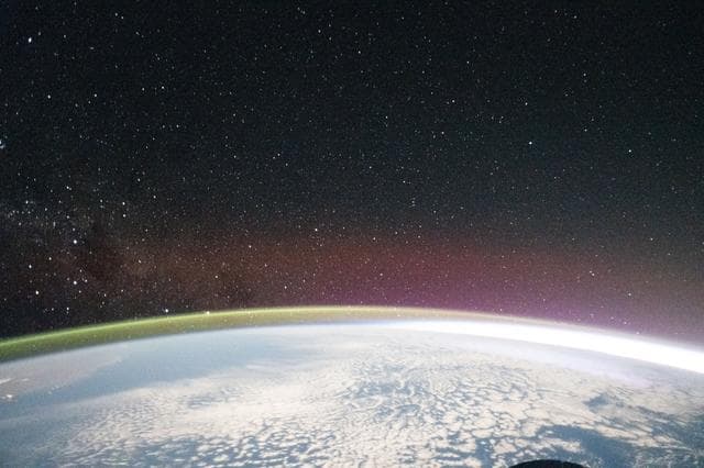 NASA image: A yellow-green airglow crowns the horizon beneath a starry sky and a faint Milky Way
