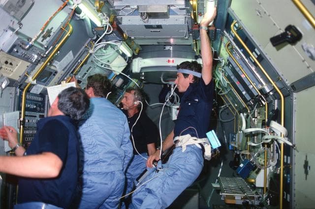 NASA image: STS-9 crewmembers gather around television monitor in Spacelab module