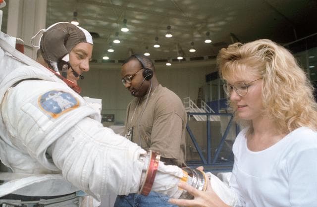NASA image: STS-53 MS Voss,in EMU, dons gloves with technicians' assistance at JSC's WETF